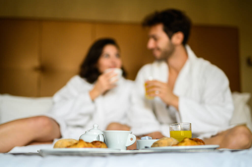 Hermosa pareja desayunar en la cama en el hotel, hombre joven y mujer en albornoces felices juntos por la mañana - Foto, imagen