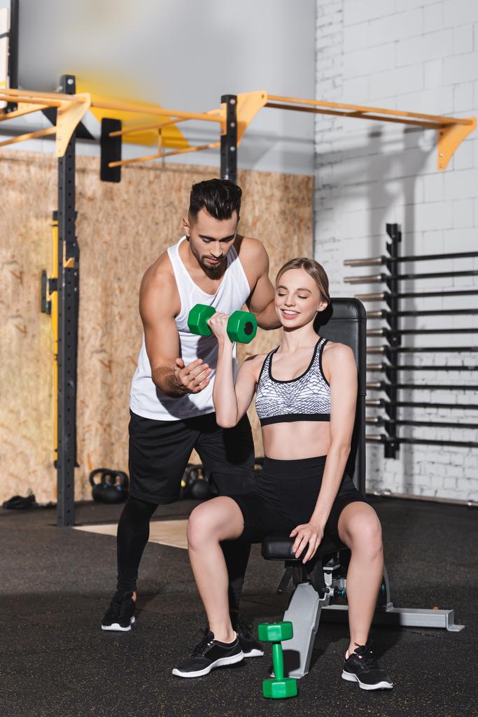 Mujer sonriente entrenando con pesas cerca del entrenador en el gimnasio  - Foto, Imagen