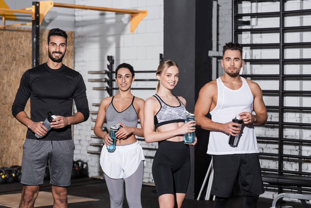 Smiling interracial sportsmen and sportswomen holding sports bottles in gym  - Photo, Image