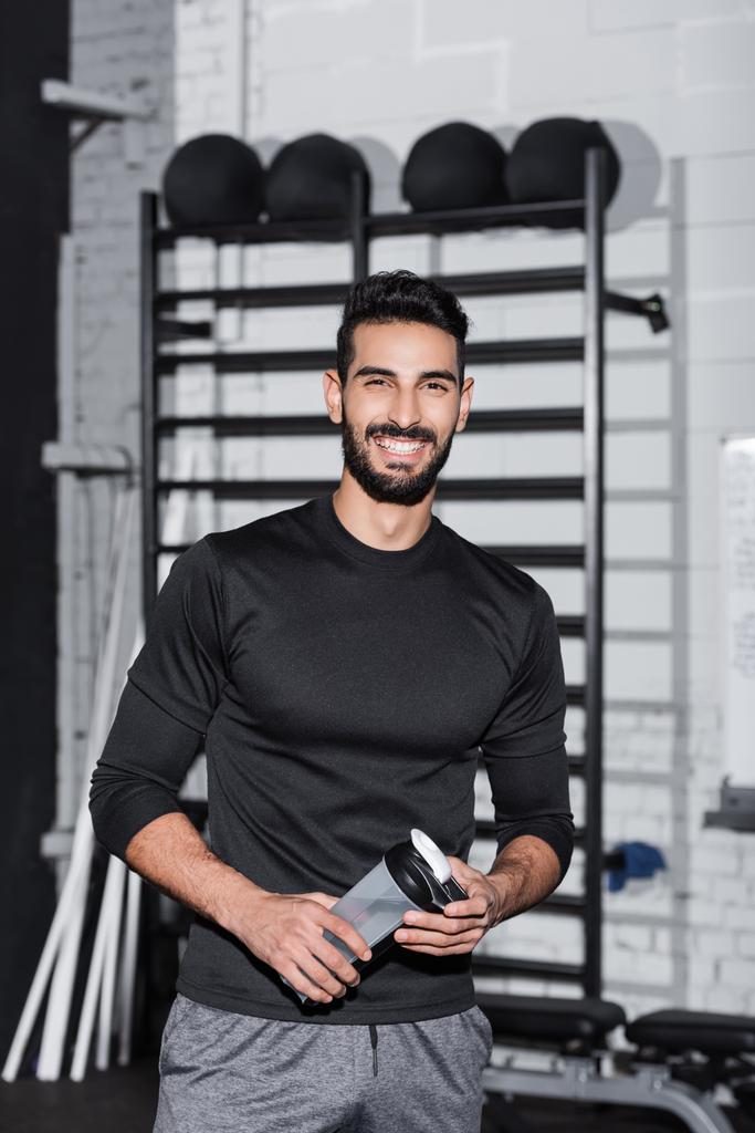 Alegre desportista árabe segurando garrafa de esportes e olhando para a câmera no ginásio  - Foto, Imagem