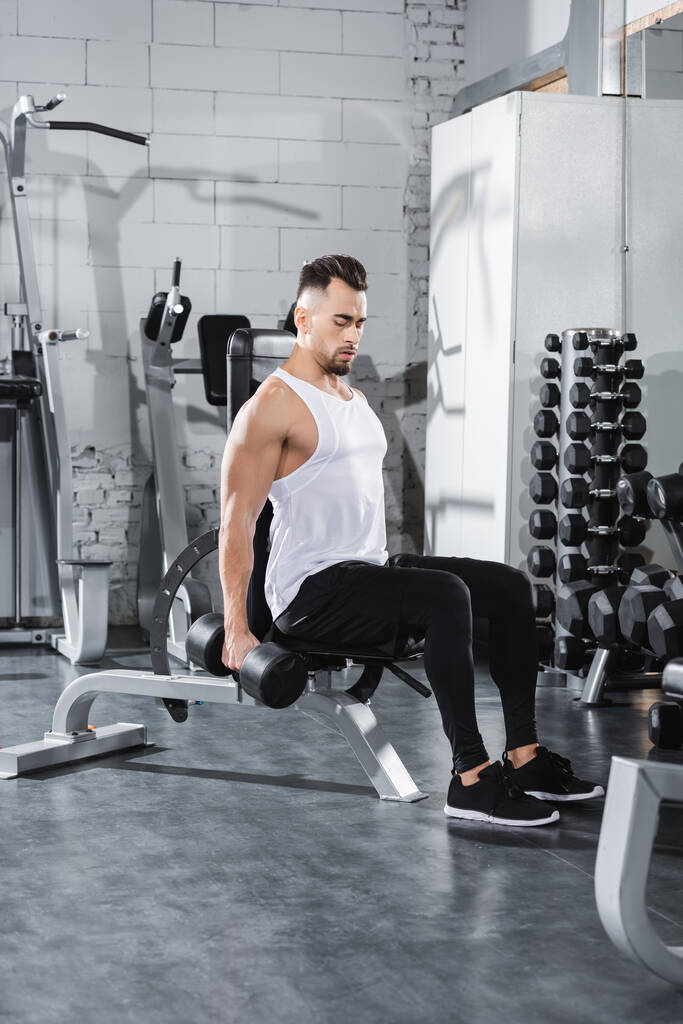 Athletic sportsman working out with dumbbell near sports equipment in gym  - Photo, Image