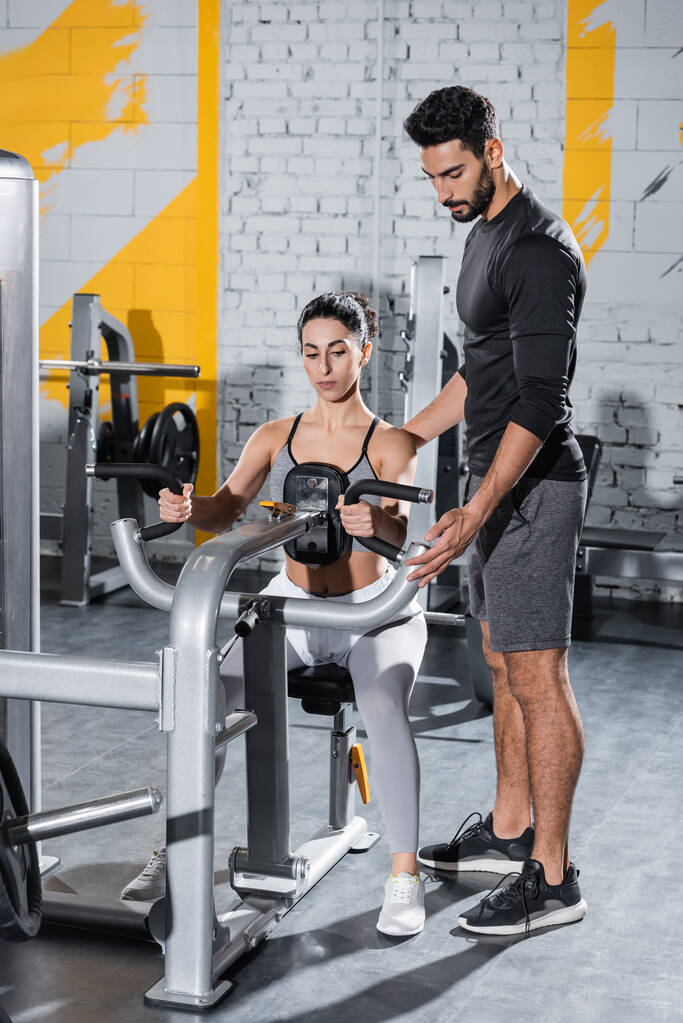 Treinador árabe de pé perto do Oriente Médio treinamento esportista na máquina ab crunch no ginásio  - Foto, Imagem