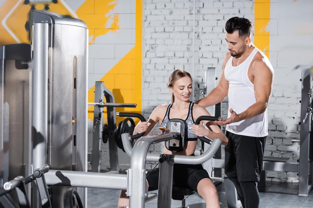 Entrenador ayudando a la deportista rubia a entrenar en la máquina ab crunch en el gimnasio  - Foto, imagen