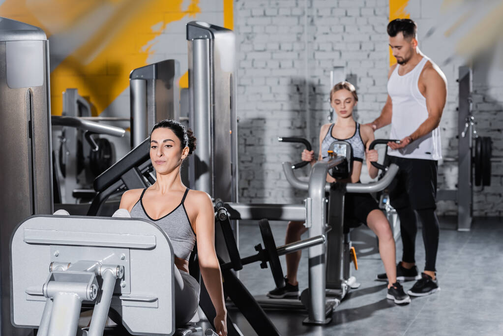 Entrenamiento de deportista de Oriente Medio en la máquina de prensa de piernas cerca de amigos borrosos en el gimnasio  - Foto, imagen
