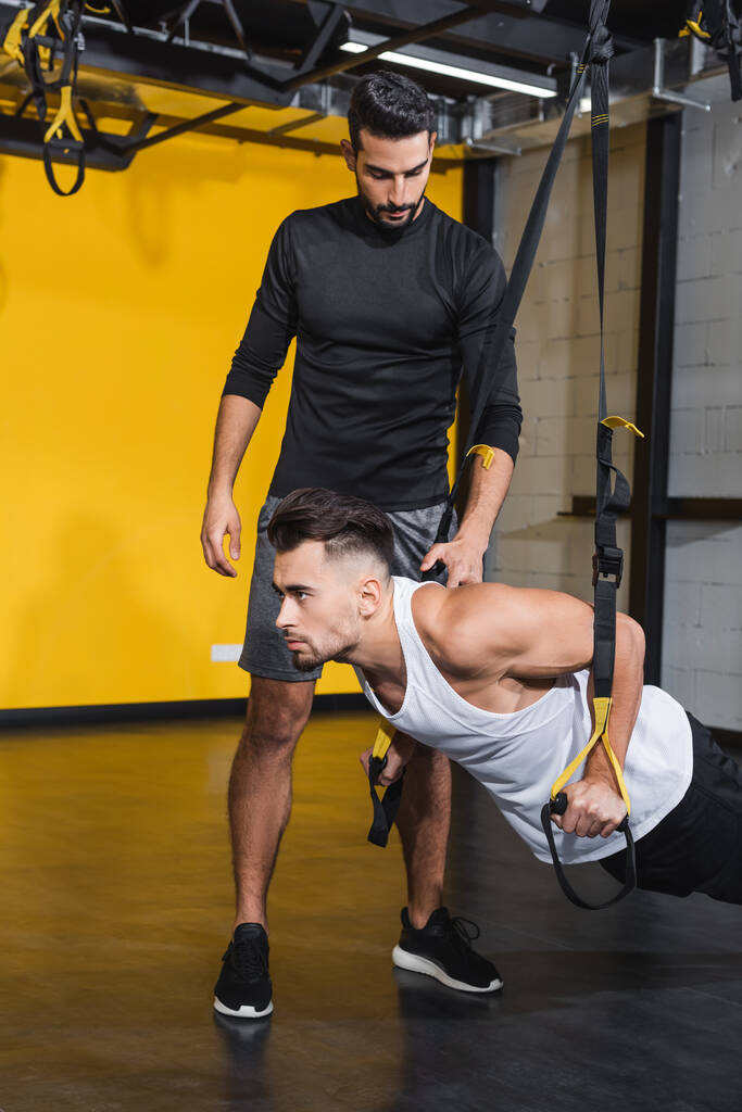 Arabischer Trainer steht neben Freund und trainiert mit Aufhängeschlaufen im Fitnessstudio  - Foto, Bild