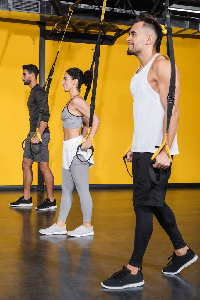 Souriant interracial personnes tirant des bretelles de suspension dans la salle de gym  - Photo, image