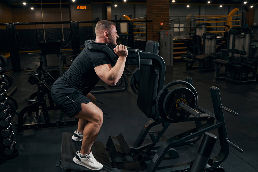 Mannelijke atleet doet squats met behulp van fitnesstoestellen - Foto, afbeelding