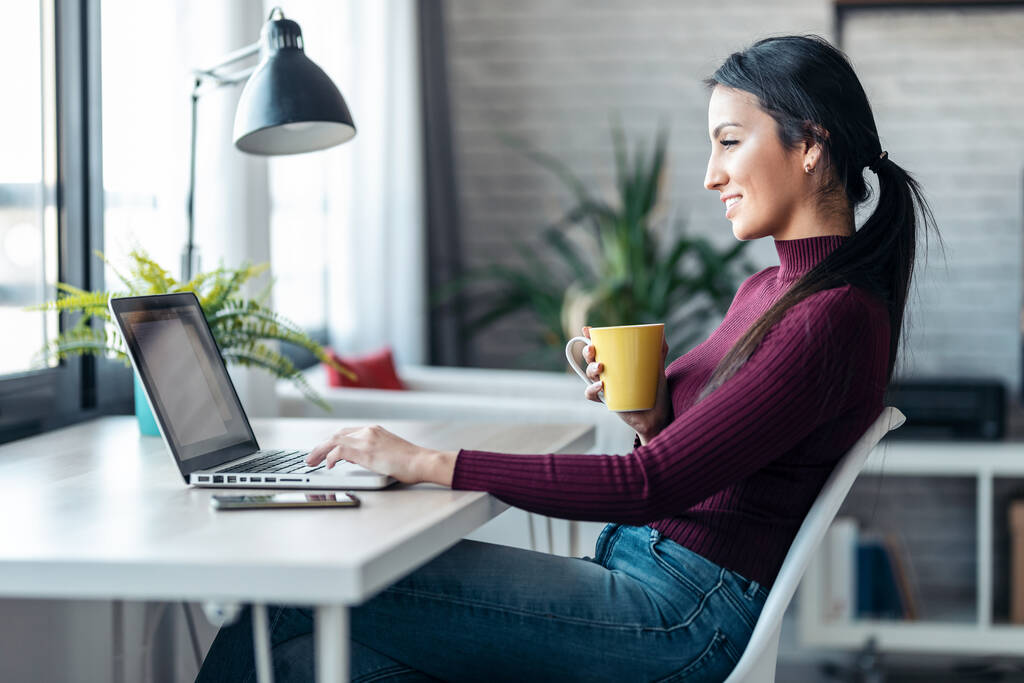 Aufnahme einer konzentrierten jungen Geschäftsfrau, die mit dem Computer arbeitet, während sie zu Hause im Büro Kaffee trinkt. - Foto, Bild