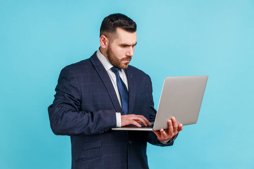 Молодой человек сконцентрировался на ношении официального костюма, держа в руках ноутбук, работая за компьютером, глядя на дисплей с серьезным выражением лица. Крытая студия снята на голубом фоне. - Фото, изображение