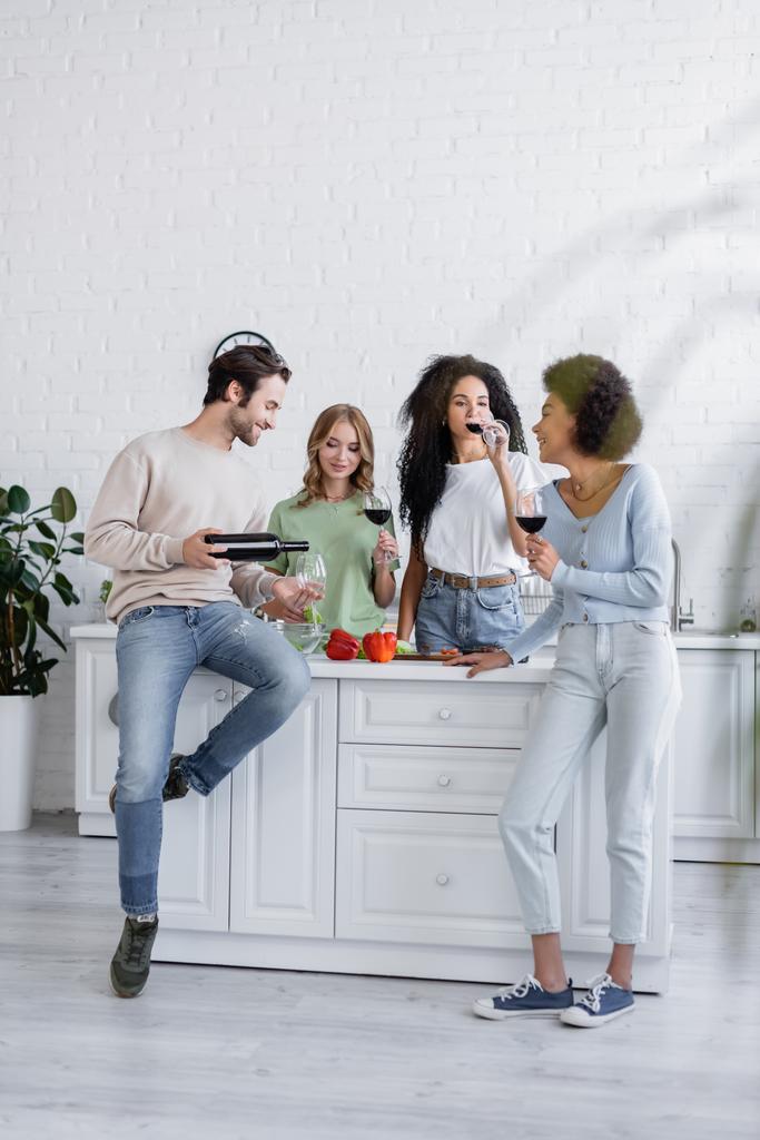 volledige lengte van vrolijke man gieten rode wijn in glas in de buurt van jonge interraciale vrouwen in keuken - Foto, afbeelding