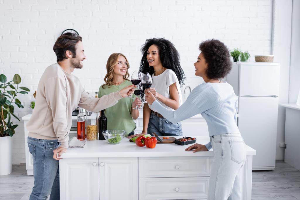 Ευτυχισμένοι διαφυλετικοί φίλοι που κλίνουν ποτήρια κόκκινο κρασί στην κουζίνα  - Φωτογραφία, εικόνα