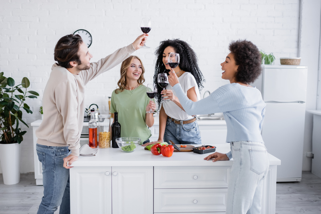 幸せな異人種間の友人がキッチンで赤ワインのグラスを持ち  - 写真・画像