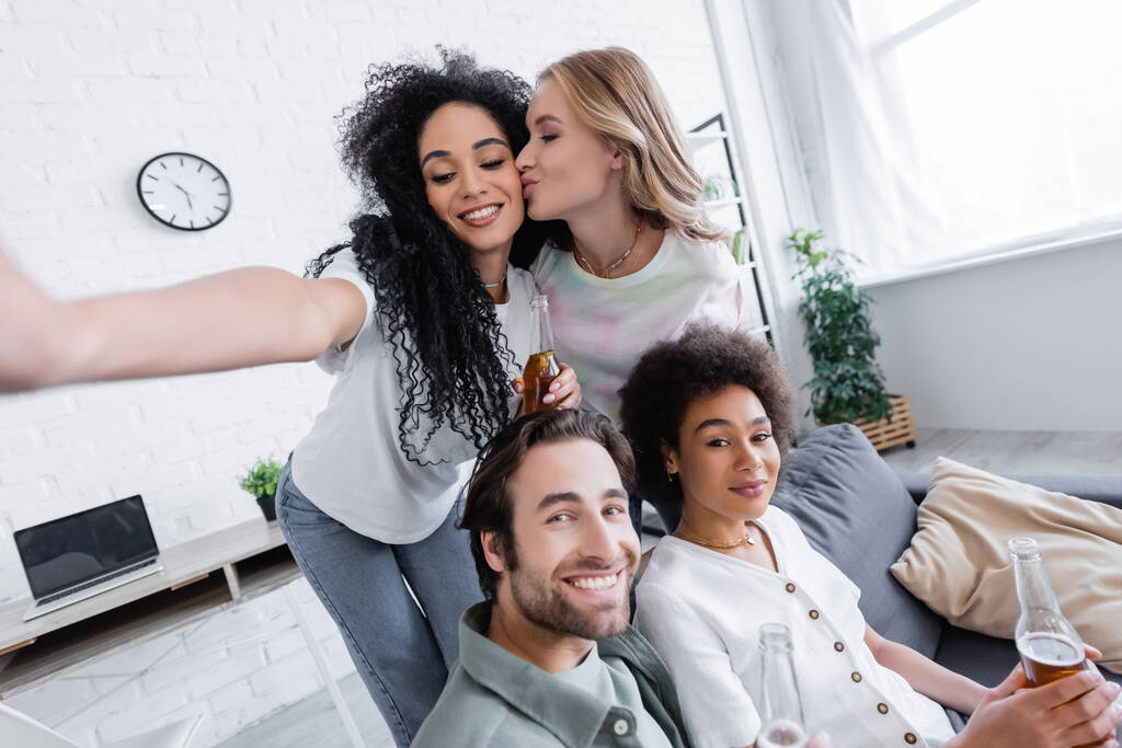 счастливая блондинка целует щеку африканской американской подружки рядом с веселыми друзьями в гостиной - Фото, изображение