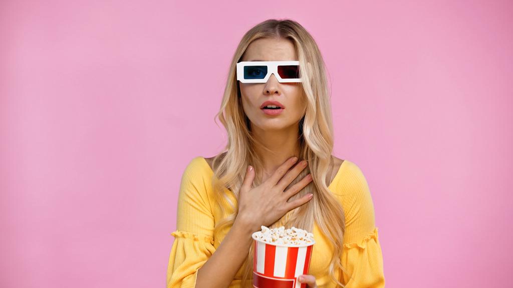 Aggódó nő 3D-s szemüvegben kezében vödör popcorn elszigetelt rózsaszín  - Fotó, kép