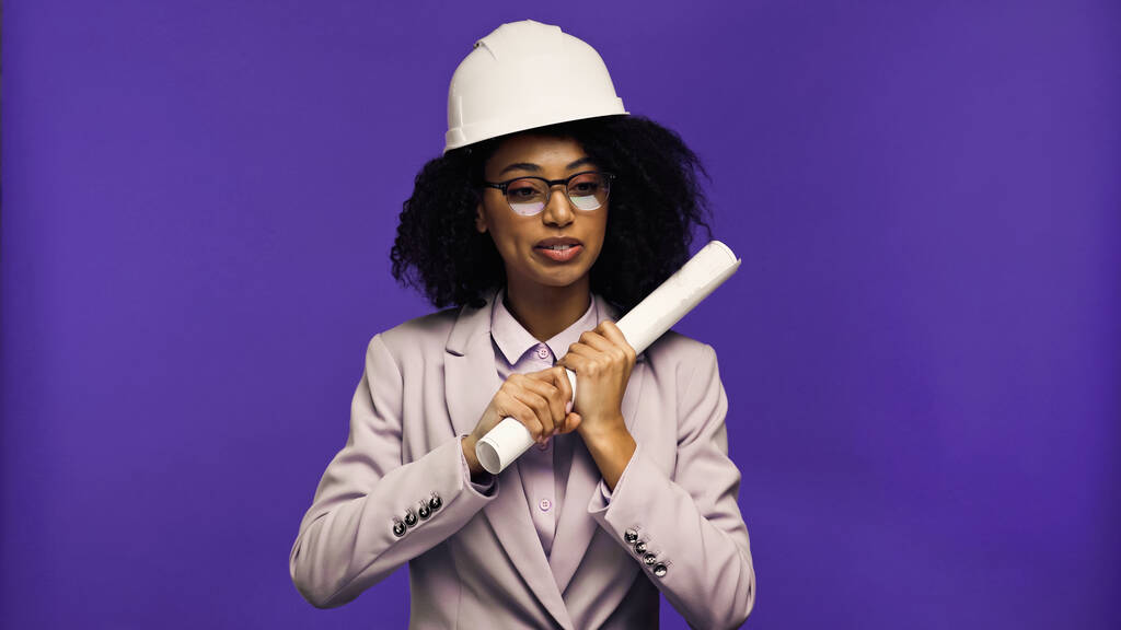 african american engineer in safety helmet holding rolled paper isolated on purple  - Photo, Image