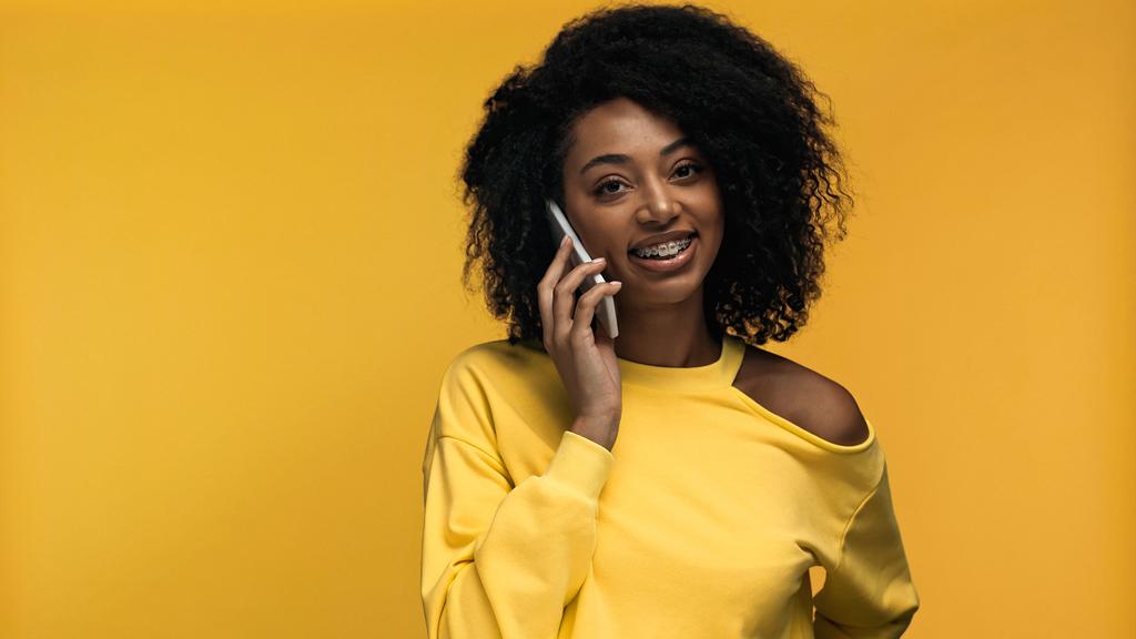 veselé africké Američanky s rovnátka mluví na smartphone izolované na žluté - Fotografie, Obrázek