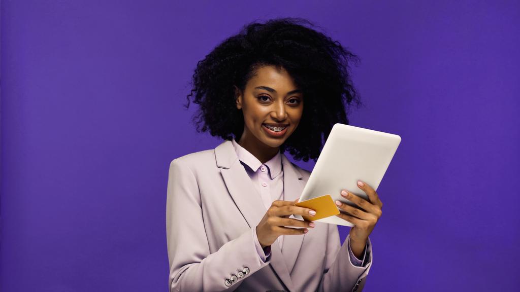 весела афроамериканка в блістері тримає цифровий планшет і кредитну картку ізольовано на фіолетовому
  - Фото, зображення