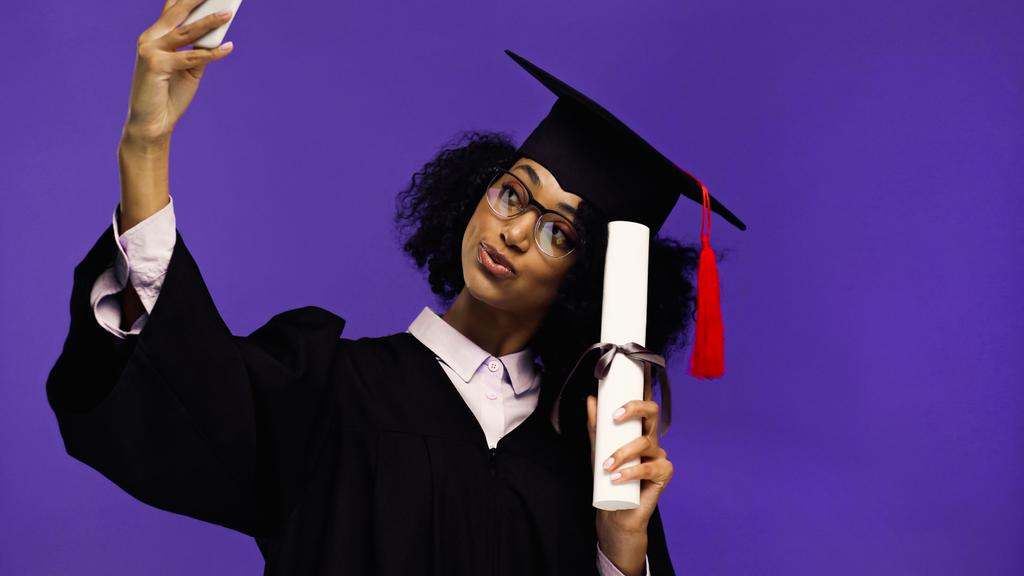 sourire étudiant afro-américain en bonnet de graduation et robe de prendre selfie avec diplôme roulé isolé sur violet  - Photo, image