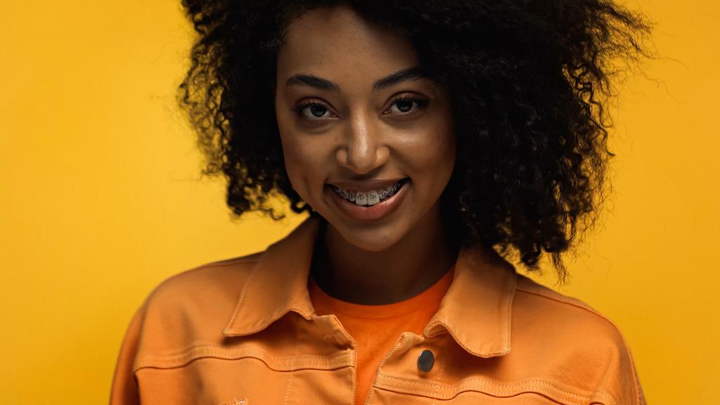 pozytywna Afroamerykanka z aparatem na zęby uśmiechnięta odizolowana na żółto - Zdjęcie, obraz