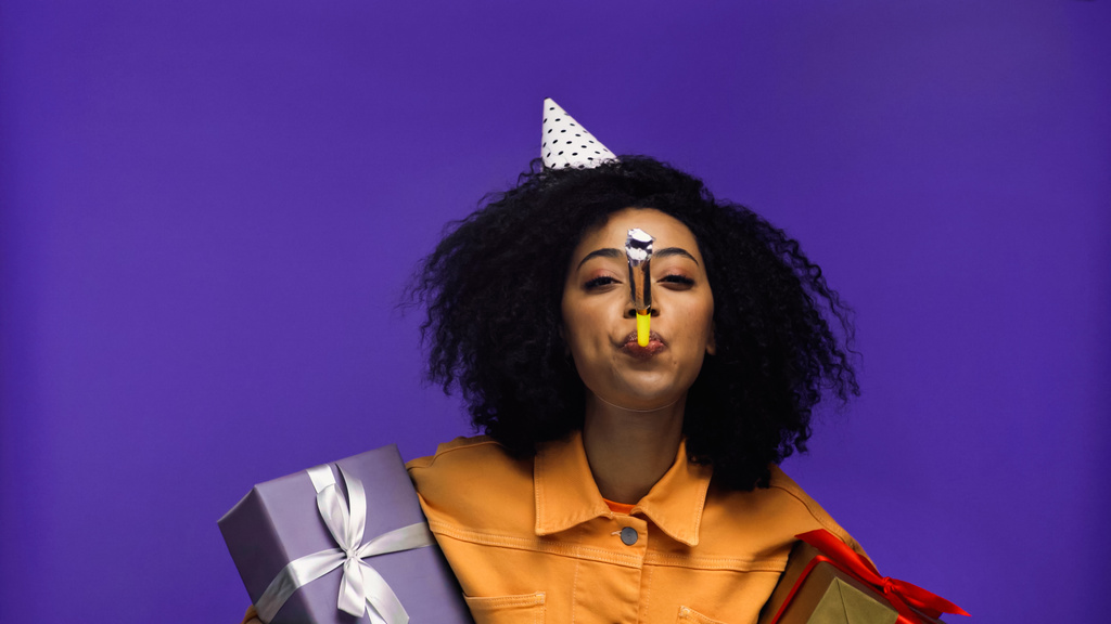 Африканская американка в шапочке для вечеринок трубит в рог вечеринки и держит подарки изолированные на фиолетовый  - Фото, изображение