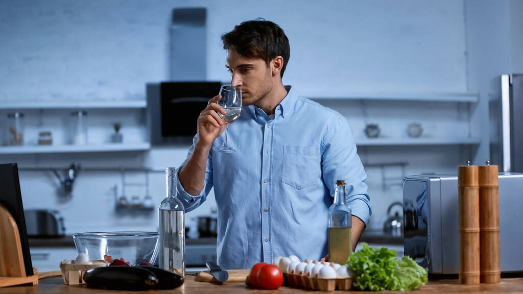 νεαρός άνδρας κρατώντας το ποτήρι και μυρίζοντας λευκό κρασί κοντά στο τραπέζι με φρέσκα συστατικά - Φωτογραφία, εικόνα