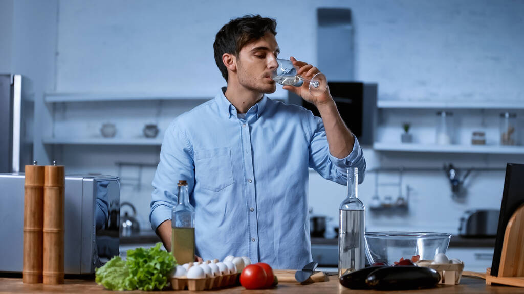 jonge man met glas en het drinken van witte wijn in de buurt van tafel met verse ingrediënten - Foto, afbeelding