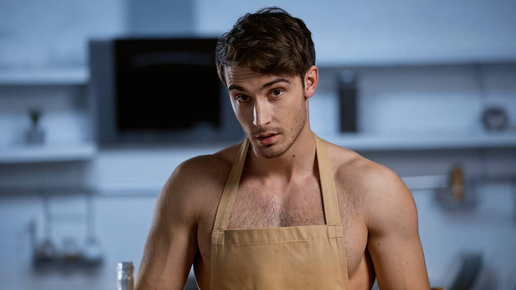 bez košile muž v béžové zástěře při pohledu na kameru v kuchyni - Fotografie, Obrázek