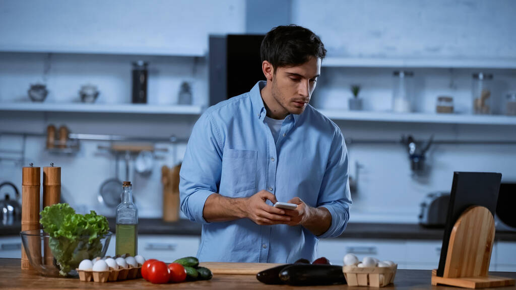 jeune homme tenant smartphone et regardant les légumes sur la table dans la cuisine  - Photo, image