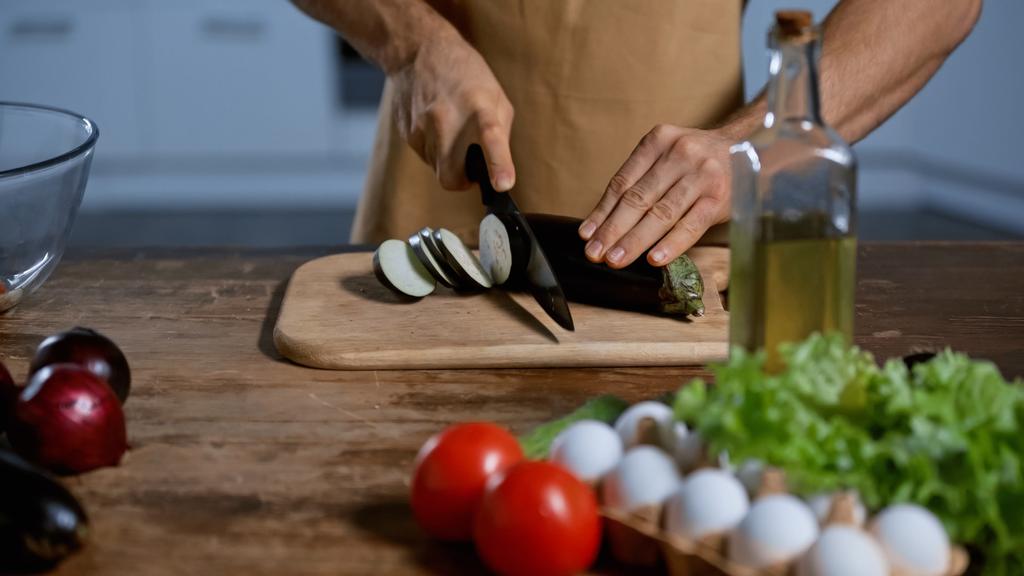zugeschnittene Ansicht eines Mannes, der Auberginen in der Nähe von Tomaten, Zwiebeln, Hühnereiern und Flaschen mit Öl schneidet - Foto, Bild