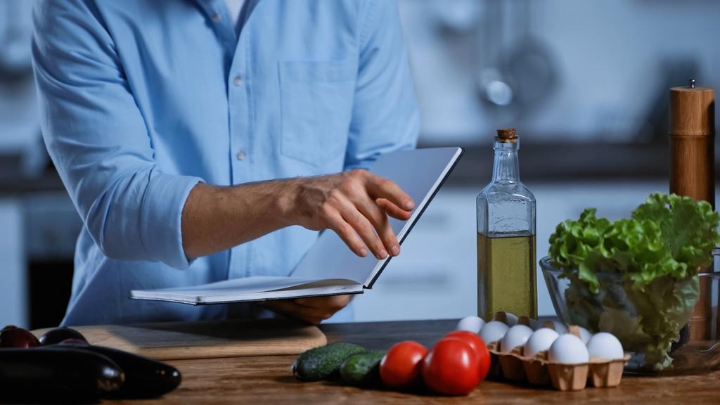 обрезанный вид человека, держащего поваренную книгу и указывающего на свежие овощи на столе - Фото, изображение
