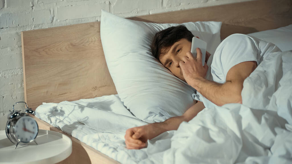 hombre hablando en el teléfono móvil mientras está acostado en la cama cerca del reloj despertador vintage - Foto, Imagen