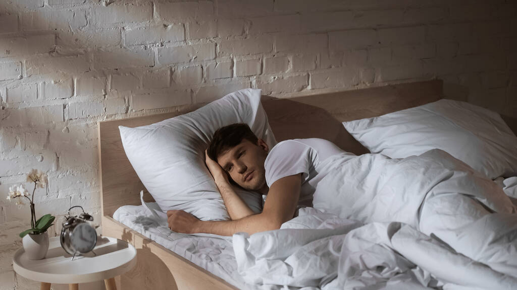 mężczyzna leżący w łóżku w pobliżu zabytkowego budzika i doniczki na stoliku nocnym - Zdjęcie, obraz