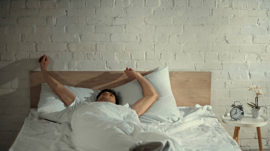 hombre estirándose en la cama cerca del despertador y orquídea en maceta en la mesita de noche - Foto, Imagen