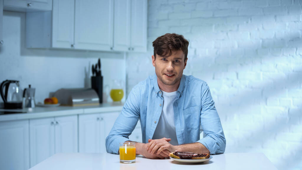 man glimlachend naar de camera terwijl hij naast de toast zit met jam en glas sinaasappelsap  - Foto, afbeelding