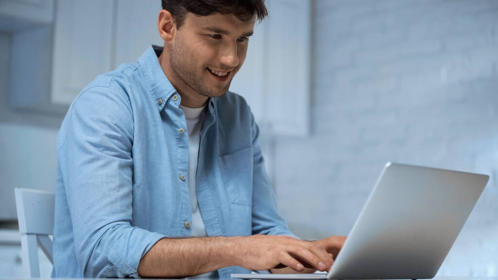 smiling freelancer in blue shirt typing on laptop in kitchen - Photo, Image