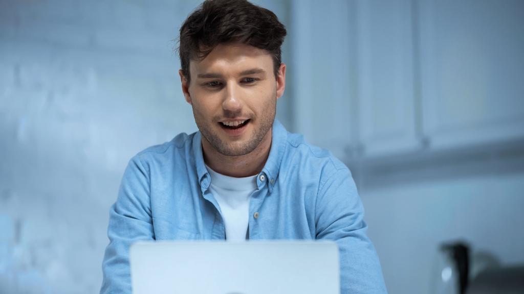sorrindo homem em camisa azul trabalhando no laptop na cozinha - Foto, Imagem