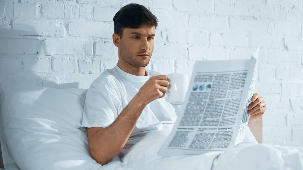 Mann mit Tasse liest Morgenzeitung, während er morgens im Bett sitzt - Foto, Bild