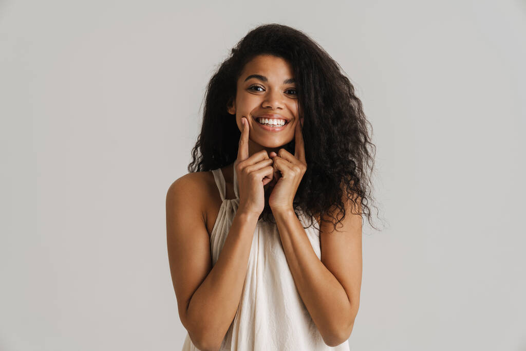 Siyah genç bir kadın gülümsüyor ve beyaz arka planda izole edilmiş yanaklarını işaret ediyor. - Fotoğraf, Görsel