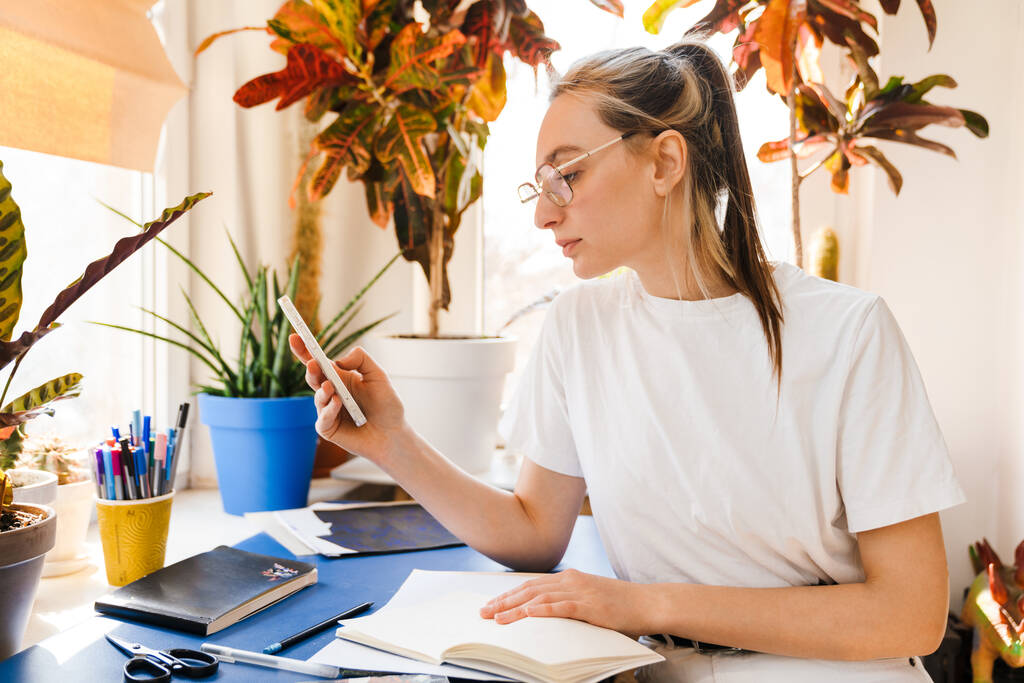 Giovane donna bianca in occhiali da vista utilizzando il telefono cellulare mentre si lavora in casa - Foto, immagini