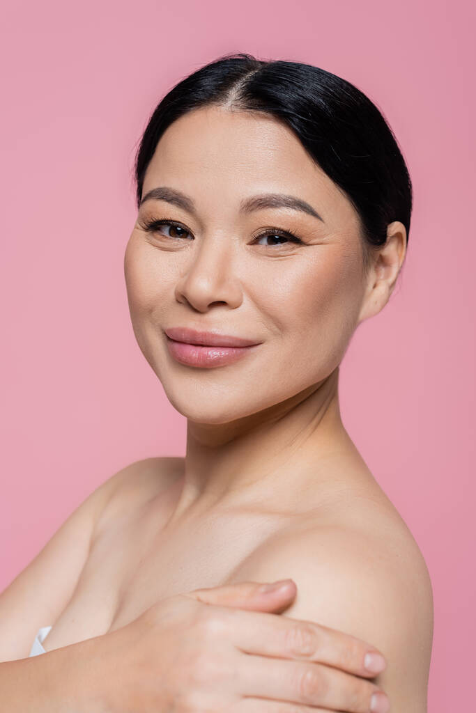 Porträt einer lächelnden asiatischen Frau mit nackten Schultern, die isoliert in die Kamera auf rosa blickt  - Foto, Bild