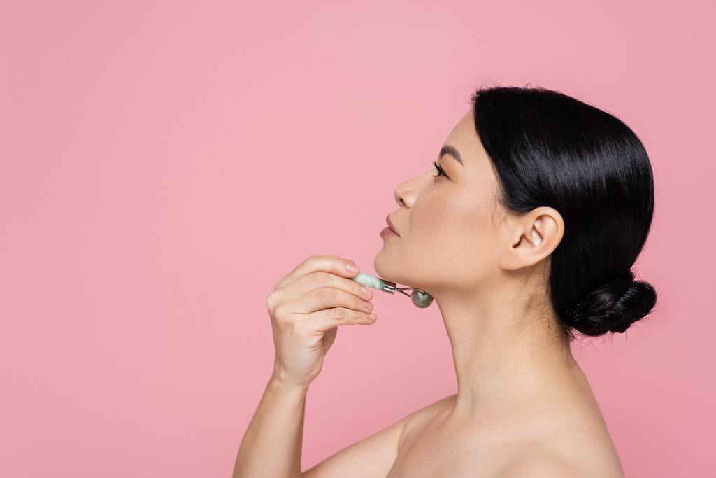 Πλευρική άποψη της γυναίκας από την Ασία με γυμνό ώμο μασάζ στο λαιμό με νεφρίτη κυλίνδρου απομονώνονται σε ροζ  - Φωτογραφία, εικόνα