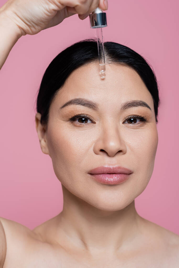 Ázsiai nő meztelen vállak alkalmazása szérum arcon izolált rózsaszín  - Fotó, kép