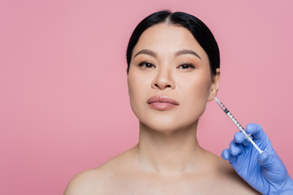 Косметолог в латексной перчатке держит шприц рядом с лицом азиатской женщины, изолированной на розовом  - Фото, изображение