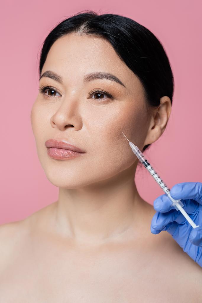 Косметолог, держащий шприц у лица азиатки, изолированной на розовом  - Фото, изображение