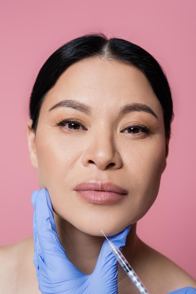 Kosmetolog drží stříkačku poblíž brady asijské ženy při pohledu na kameru izolované na růžové  - Fotografie, Obrázek