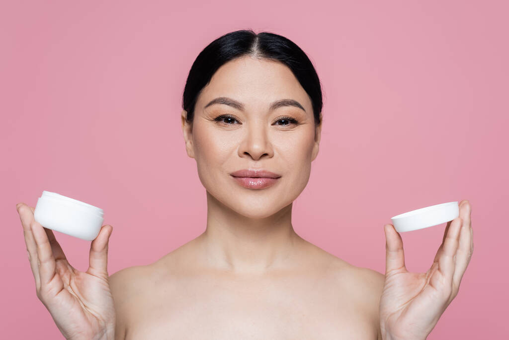 Asian woman with naked shoulders holding container with cosmetic cream isolated on pink  - Photo, Image