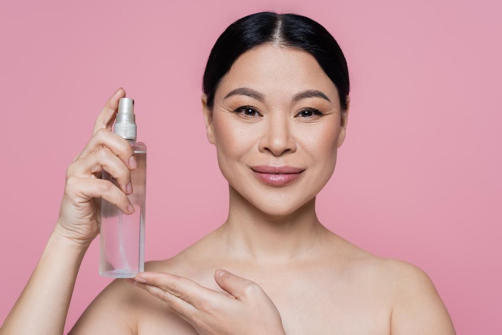 Smiling asian woman with naked shoulders holding bottle with thermal water isolated on pink  - Photo, Image