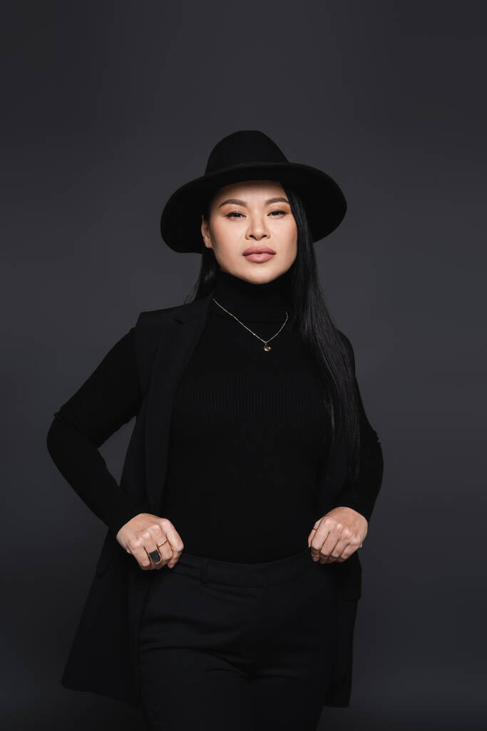 Stilvolle asiatische Frau mit Fedora-Hut und Rollkragen schaut isoliert in die Kamera auf dunkelgrau - Foto, Bild