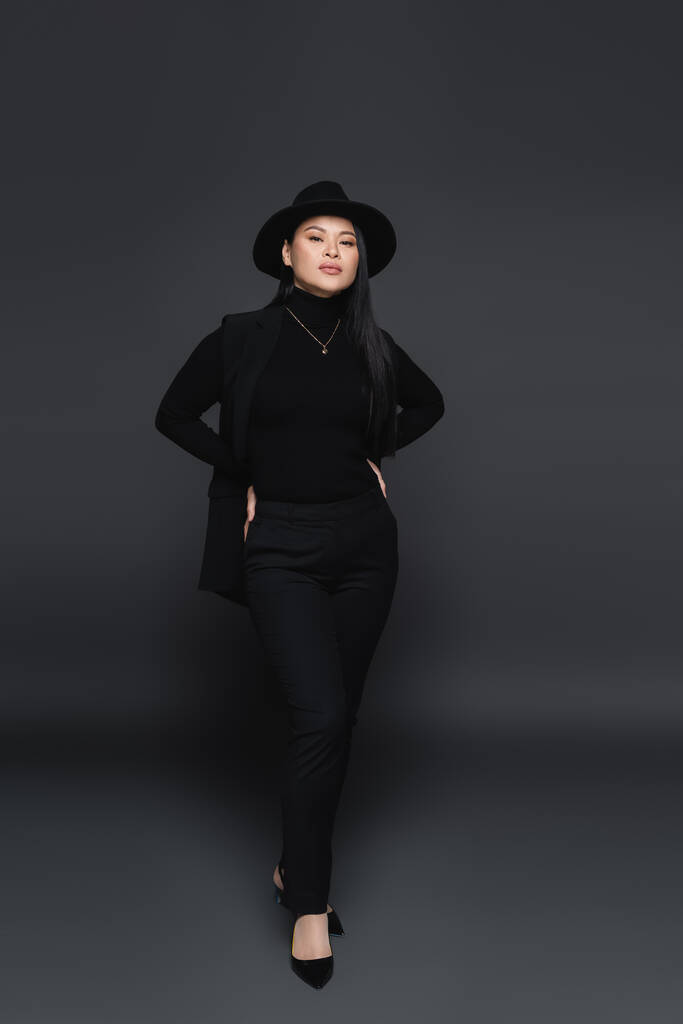 Plná délka stylové asijské ženy v klobouku fedora držet ruce v bok na tmavě šedém pozadí  - Fotografie, Obrázek