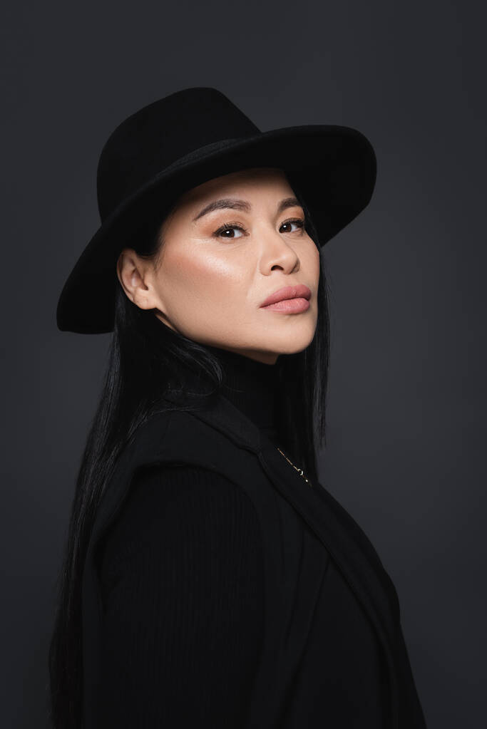 Porträt eines asiatischen Models in Jacke und Fedora-Hut, das isoliert auf dunkelgrau in die Kamera blickt - Foto, Bild